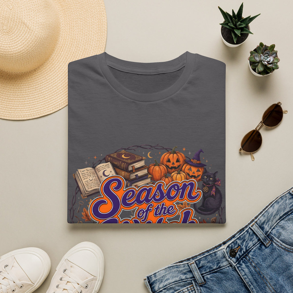 Folded charcoal grey t-shirt with Season of the Witch lettering with pumpkins, spellbooks and a witch hat, styled flat lay.