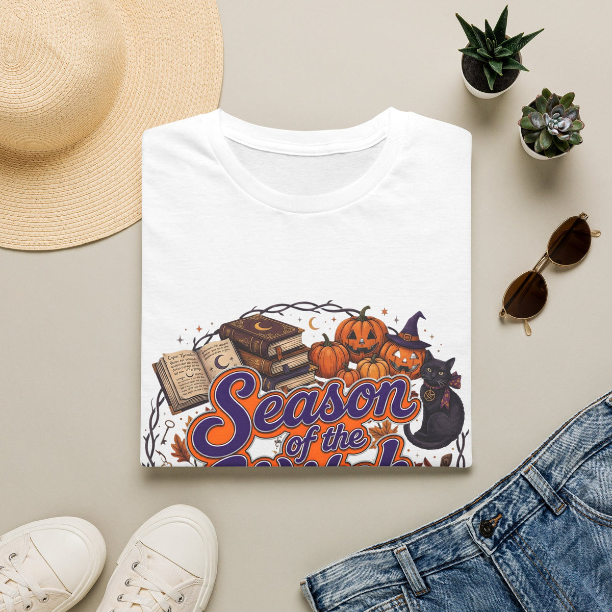 Folded white t-shirt with Season of the Witch lettering with pumpkins, spellbooks and a witch hat, styled flat lay.