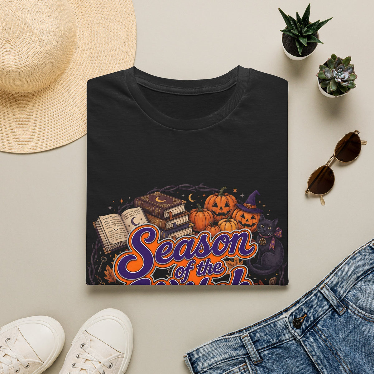 Folded black t-shirt with Season of the Witch lettering with pumpkins, spellbooks and a witch hat, styled flat lay.