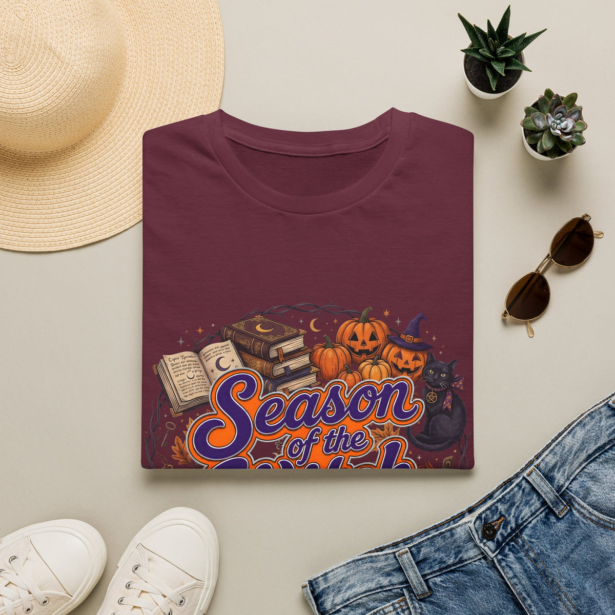 Folded burgundy t-shirt with Season of the Witch lettering with pumpkins, spellbooks and a witch hat, styled flat lay.